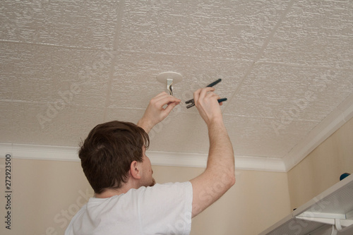 Молодой мужчина в белой футболке с отверткой вешает светильник на потолок