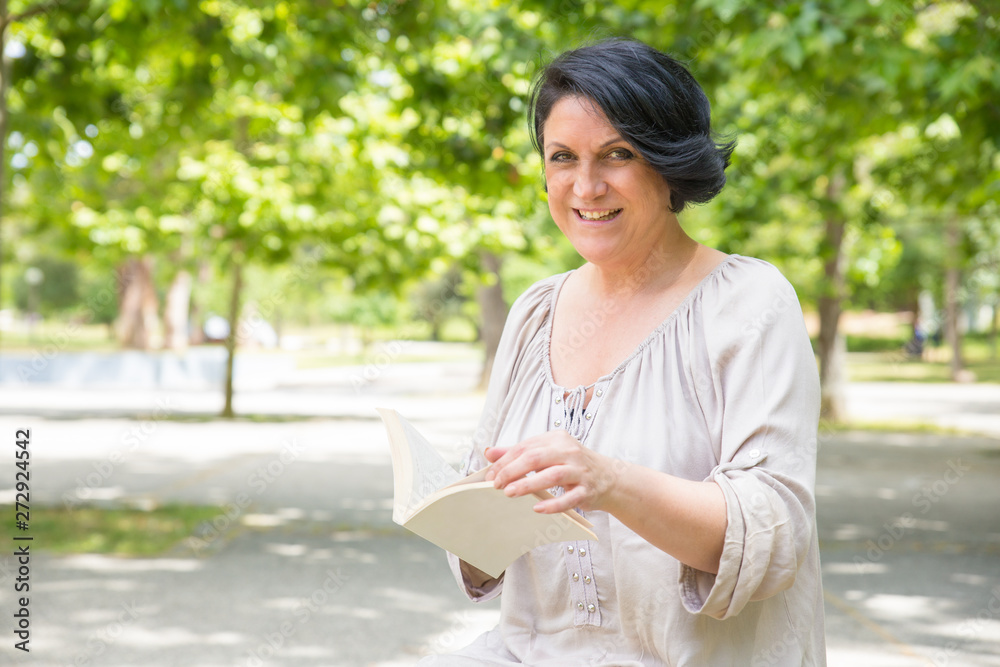 Happy cheerful woman with book relaxing in park. Middle aged black haired lady turning pages of book and smiling at camera. Leisure time in park concept Happy cheerful woman with book relaxing in park. Middle aged black haired lady turning pages of book and smiling at camera. Leisure time in park concept