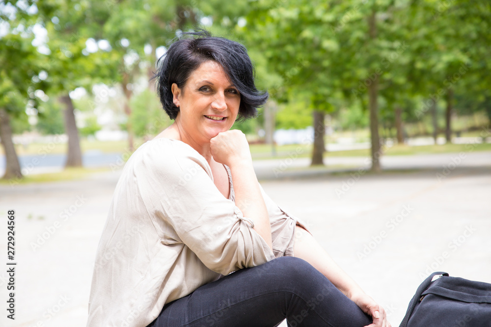 Happy confident woman relaxing in park. Middle aged black haired lady sitting against green trees and smiling at camera. Woman outdoors concept Happy confident woman relaxing in park. Middle aged black haired lady sitting against green trees and smiling at camera. Woman outdoors concept