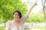 Joyful excited woman standing outdoors against green trees and pointing finger up. Middle aged black haired lady raising hand and smiling at camera. Pointing up concept
