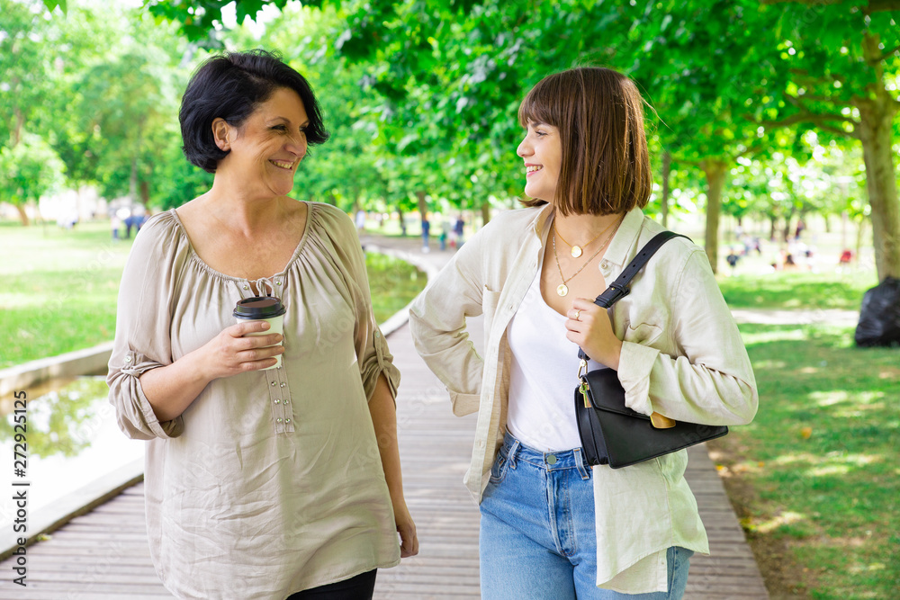 Happy young woman and her mother chatting and walking in park. Middle-aged lady and her daughter walking on boardwalk and relaxing with trees in background. Family and nature concept. Front view. Happy young woman and her mother chatting and walking in park. Middle-aged lady and her daughter walking on boardwalk and relaxing with trees in background. Family and nature concept. Front view.