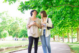 Positive young woman and her mother chatting and walking in park. Middle-aged lady and her daughter walking on boardwalk and relaxing with trees in background. Family and nature concept. Front view.
