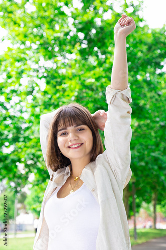 Happy pretty young woman raising arms in city park. Lady standing and looking at camera with green trees in background. Leisure and summer concept. Front view.