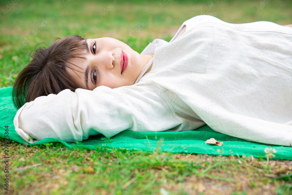 Smiling pretty young woman lying and relaxing on lawn. Lady looking at camera and resting on blanket on ground. Relaxation and nature concept. Side view. Smiling pretty young woman lying and relaxing on lawn. Lady looking at camera and resting on blanket on ground. Relaxation and nature concept. Side view.