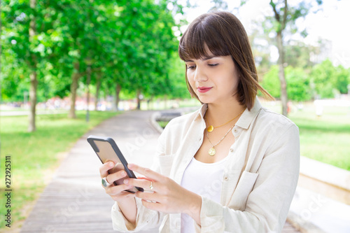 Serious young woman using smartphone in city park. Pretty lady wearing casual shirt and standing with trees and walkway in background. Communication and nature concept.