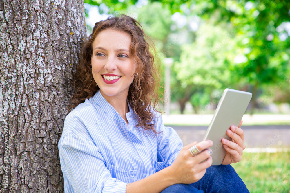 Happy beautiful woman using mobile internet on tablet. Positive pretty girl sitting by tree in summer park. Modern device concept Happy beautiful woman using mobile internet on tablet. Positive pretty girl sitting by tree in summer park. Modern device concept