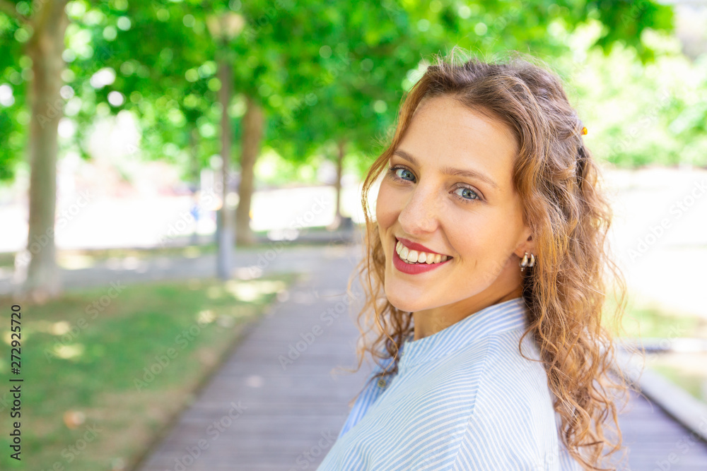 Happy wavy-haired girl smiling at camera outdoors. Portrait of cheerful young woman with earrings walking along park pathway. Summer concept 
