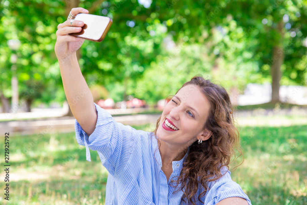 Jolly charming lady winking while taking selfie in park. Happy young woman with wavy hair resting alone outdoors. Social media concept Jolly charming lady winking while taking selfie in park. Happy young woman with wavy hair resting alone outdoors. Social media concept