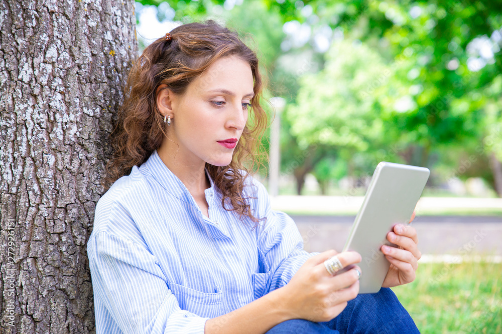 Serious young woman reading article on gadget outdoors. Concentrated wavy-haired girl leaning on tree while using tablet. Technology concept Serious young woman reading article on gadget outdoors. Concentrated wavy-haired girl leaning on tree while using tablet. Technology concept
