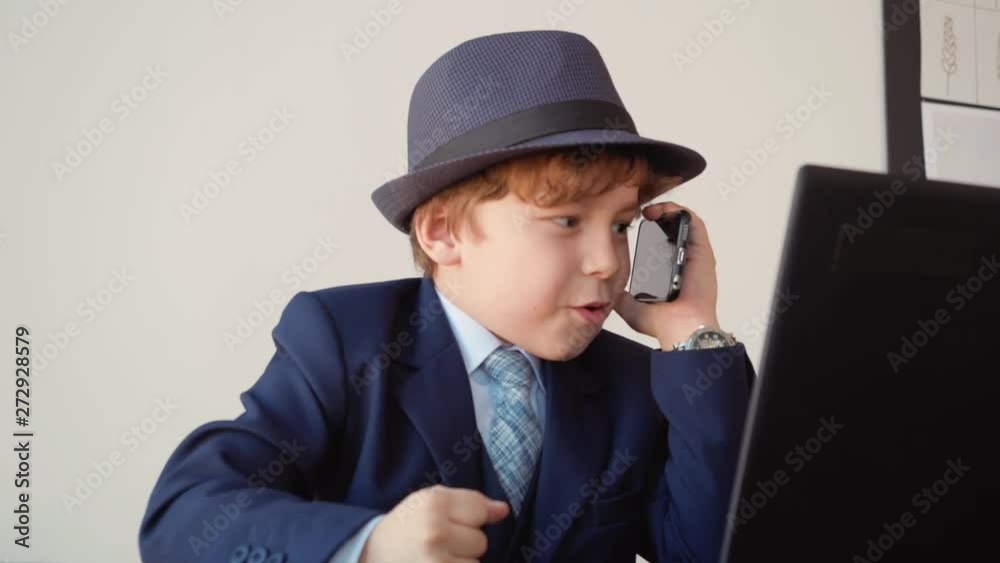 Portrait of little angry boy looks like a boss is talking mobile phone in his office sitting at table. He is wearing suit and hat. Manager in office Portrait of little angry boy looks like a boss is talking mobile phone in his office sitting at table. He is wearing suit and hat. Manager in office
