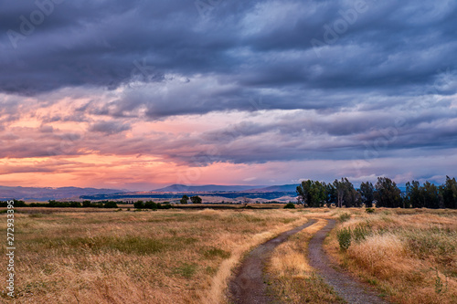 Landscape with dirt road ruts passing through a rural country field with a dramatic, colorful sunset in the background.