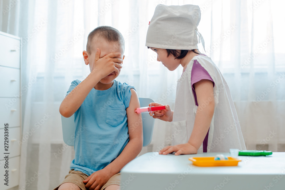Fototapeta premium Children play in the hospital. The girl is dressed in a doctor 4 years old makes an injection to a boy 6 years old with a toy syringe in a room in a kindergarten.