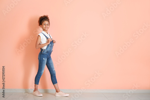 Stylish African-American girl in jeans overall near color wall