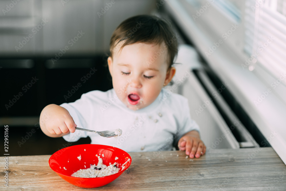Funny little girl in the kitchen during the day eating oatmeal porridge from a red plate all dirty Funny little girl in the kitchen during the day eating oatmeal porridge from a red plate all dirty