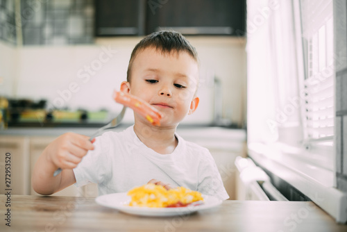 a child in a t-shirt in the kitchen eating a sausage and an omelet with a fork is very appetizing