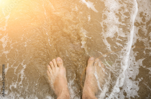 Man is relaxing barefoot at the beach. Guy's legs in waves at the ocean side. Coastline in the clouds and sun. Summer vacation. Travel concept.