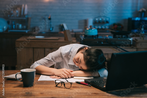 Sleepy exhausted asian woman employee working at wooden kitchen desk with laptop computer. lady eyes are closing and about to fall asleep. sleep deprivation and overtime working concept in midnight. photo