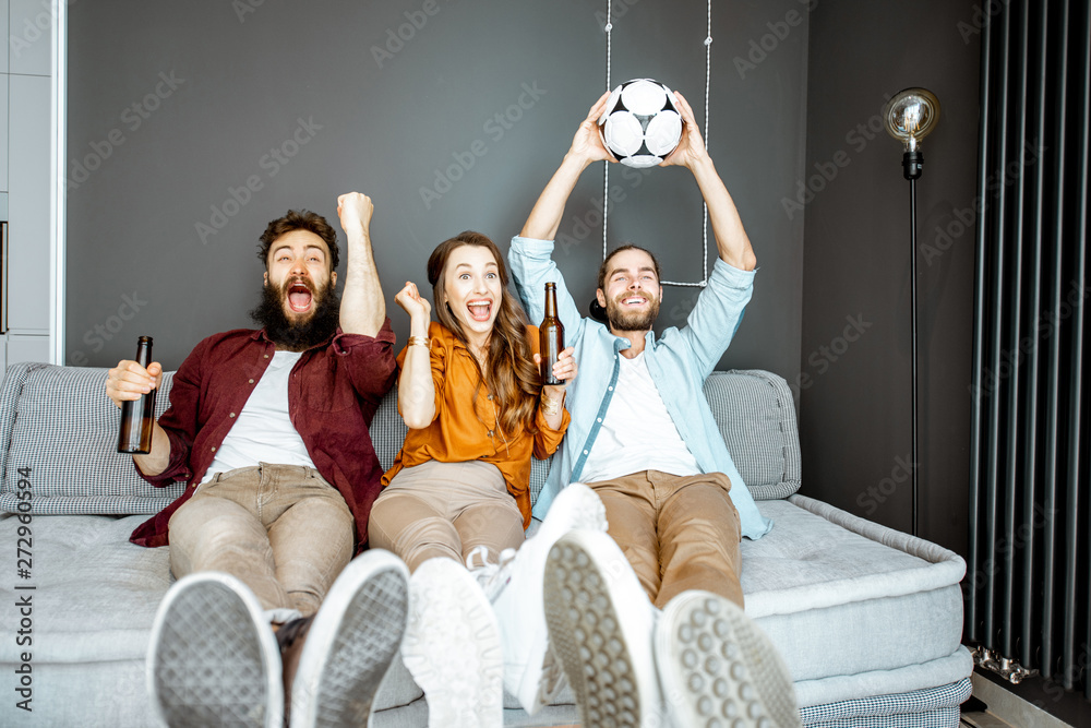 Young friends watching football match, cheering up for a favorite team while sitting together with drinks and ball on the couch at home