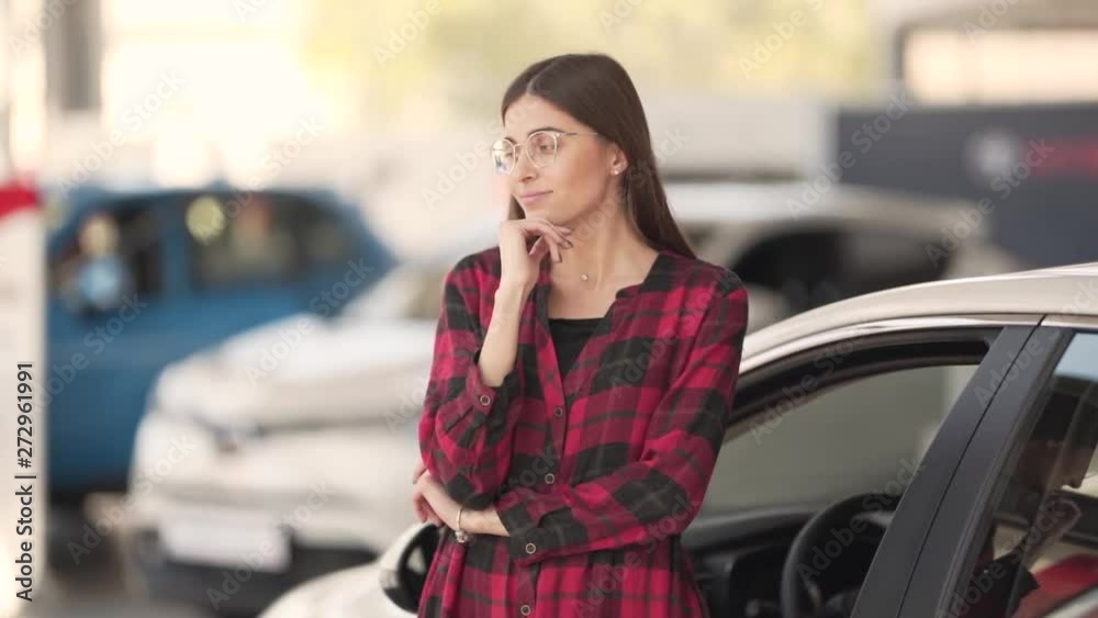 Young Happy Caucasian Female With Glasses Standing By New White Car in Indoor Showroom Thinking About Something. Blurred Background Young Happy Caucasian Female With Glasses Standing By New White Car in Indoor Showroom Thinking About Something. Blurred Background
