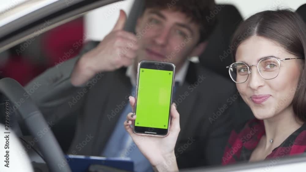 Young woman sits in her new car with a salesman and smiles at the camera while holding a cellphone with a greenscreen display. Young woman sits in her new car with a salesman and smiles at the camera while holding a cellphone with a greenscreen display.