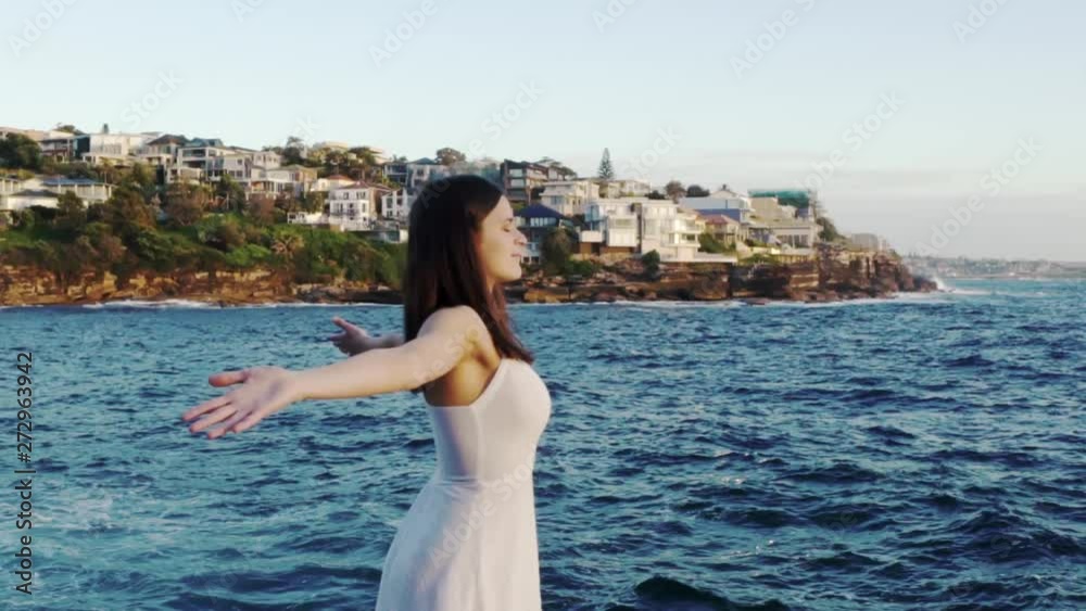 Slowmotion of Young Caucasian Female in White Summer Dress Standing On Cliff By Sea Watching Sunset Waves and Spreads Hands Slowmotion of Young Caucasian Female in White Summer Dress Standing On Cliff By Sea Watching Sunset Waves and Spreads Hands