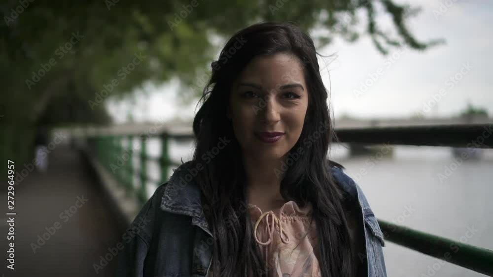 Portrait of an attractive latina tourist with black wavy hair and a jean jacket posing in a park in London with a view of Putney bridge behind her Portrait of an attractive latina tourist with black wavy hair and a jean jacket posing in a park in London with a view of Putney bridge behind her
