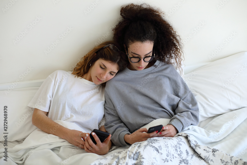 Young relaxed lesbian couple in bed texting on smartphones. - obrazy, fototapety, plakaty 