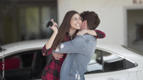 Cute young couple is standing in front of car, holding a key and greeting each other with a purchase. Thet are laughing and hugging. photo