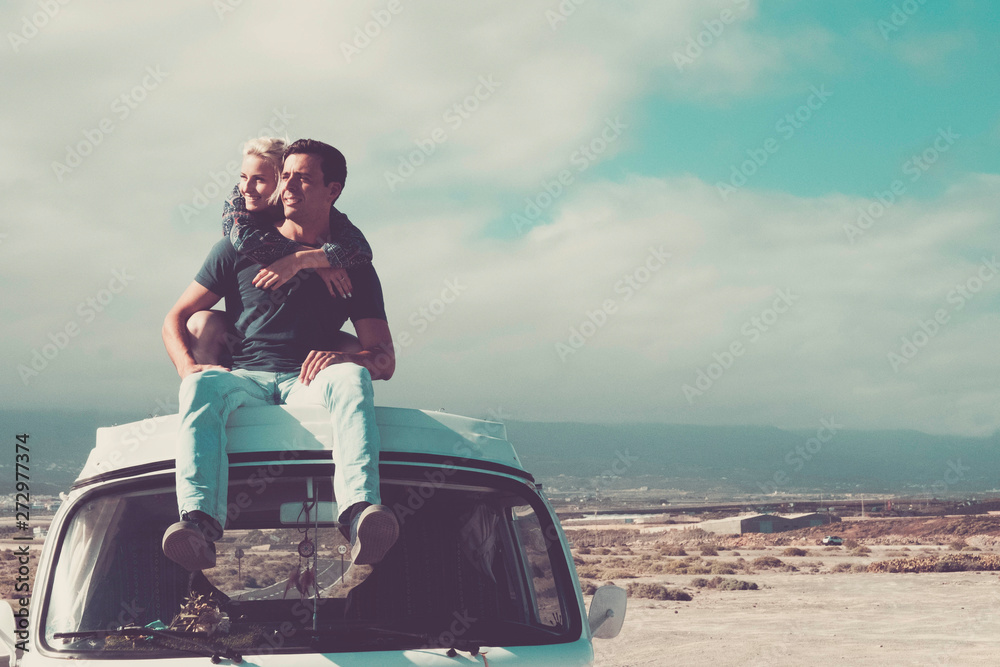 Travel people concept with young couple of man and woman sitting on the roof of old vintage van - love and relationship for travelers - beautiful landscape background - obrazy, fototapety, plakaty 