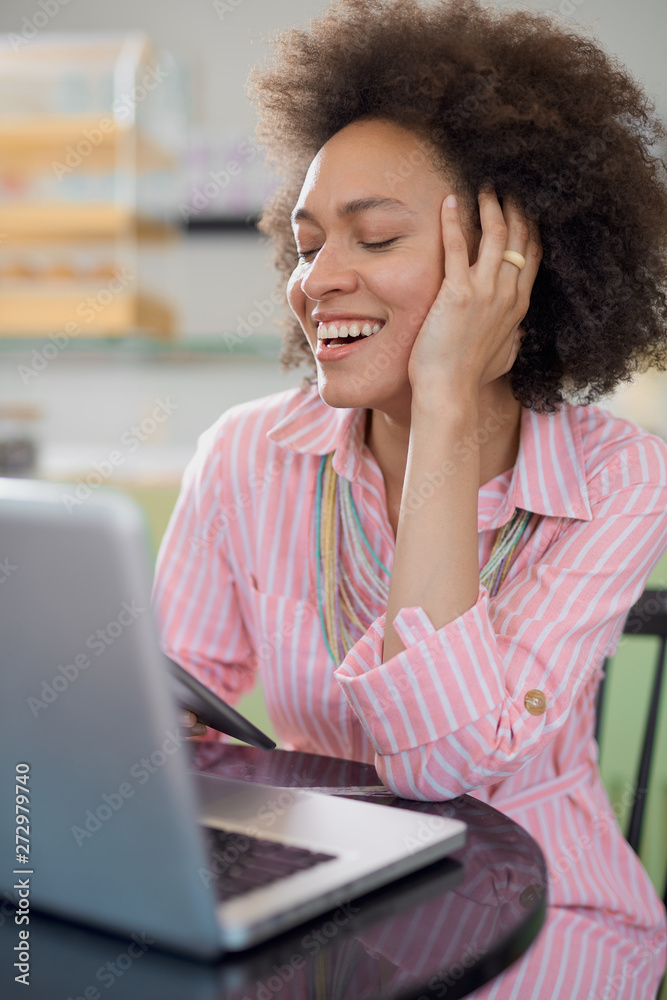 Fototapeta premium Attractive mixed race woman in striped pink dress sitting in cafe and using tablet.