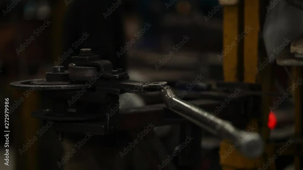 Close up of an idle metal bending machine with a worker hammering a metal bar in the background. Close up of an idle metal bending machine with a worker hammering a metal bar in the background.