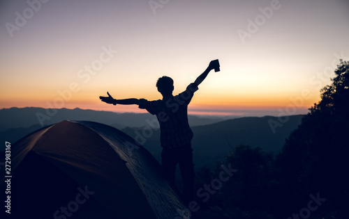 Happy hiker man holding coffee cup near camping tent on mountains at sunset background. travel concept.