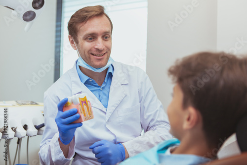 Mature cheerful male dentist educationg his young patient, showing tooth model, explaining teeth care. Professional dentist talking to a little boy photo