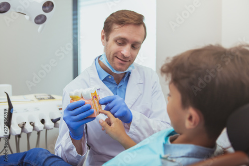 Experienced friendly dentist working at his clinic  talking to the patient  showing tooth model.Cheerful male dentist education child about dental hygiene