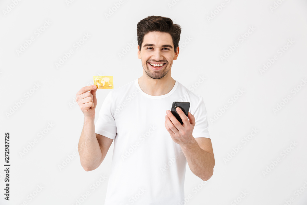 Excited happy young man posing isolated over white wall background using mobile phone holding credit card. Excited happy young man posing isolated over white wall background using mobile phone holding credit card.