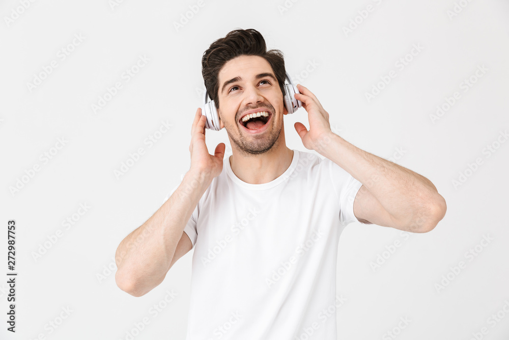 Emotional excited young man posing isolated over white wall background listening music with headphones. Emotional excited young man posing isolated over white wall background listening music with headphones.