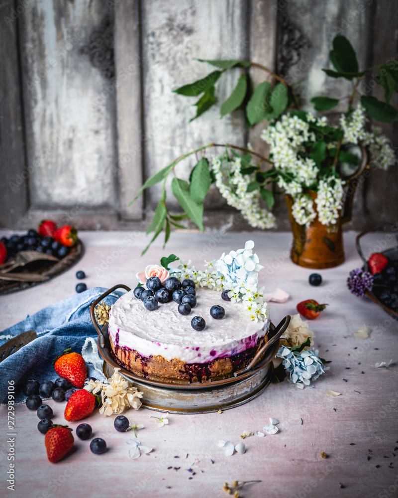 cheesecake with fresh blueberries on metal tray among fresh berries, flowers and textile on shabby gray background