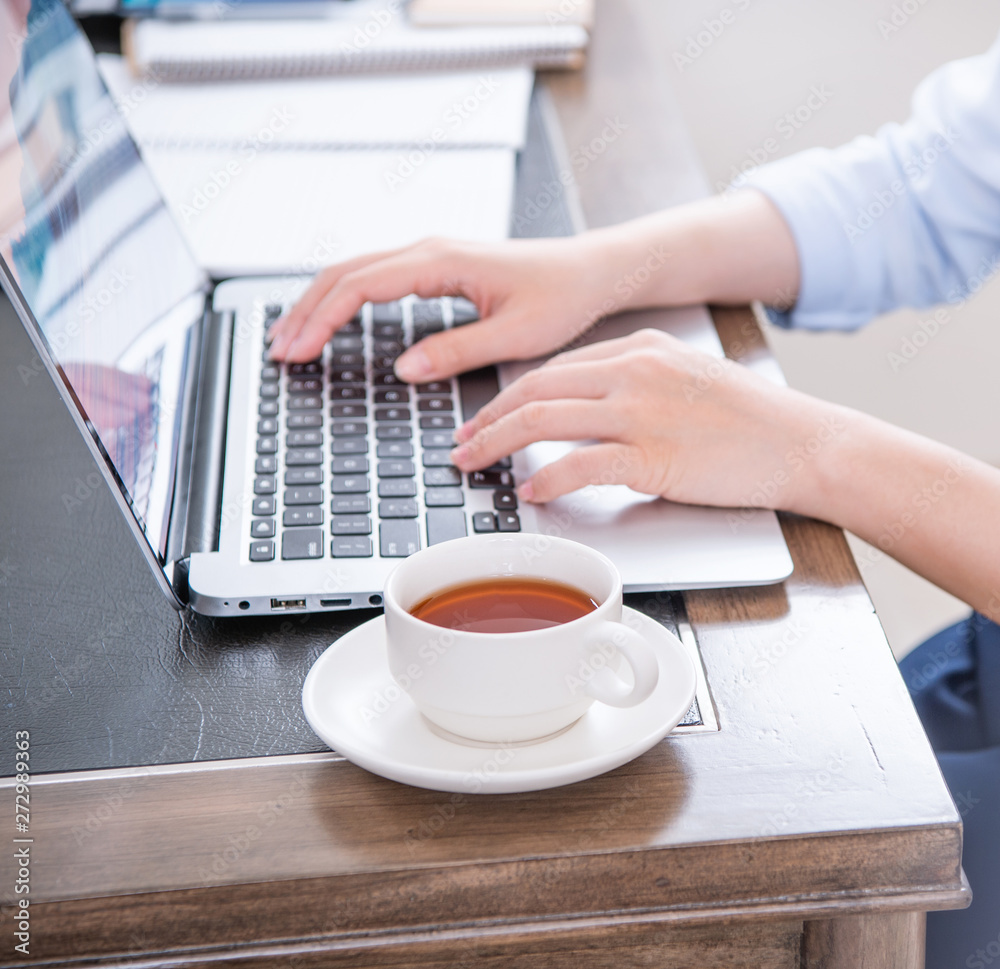 Business concept. Woman in blue shirt typing on computer with coffee on office table, backlighting, sun glare effect, close up, side view, copy space Business concept. Woman in blue shirt typing on computer with coffee on office table, backlighting, sun glare effect, close up, side view, copy space