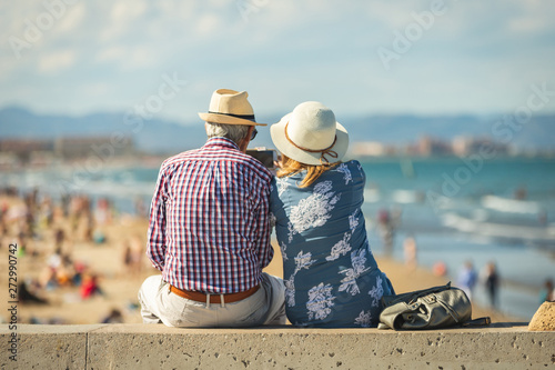 Loving the elderly couple sitting on the wall facing the beach, watching and taking pictures of the landscape on a romantic trip photo