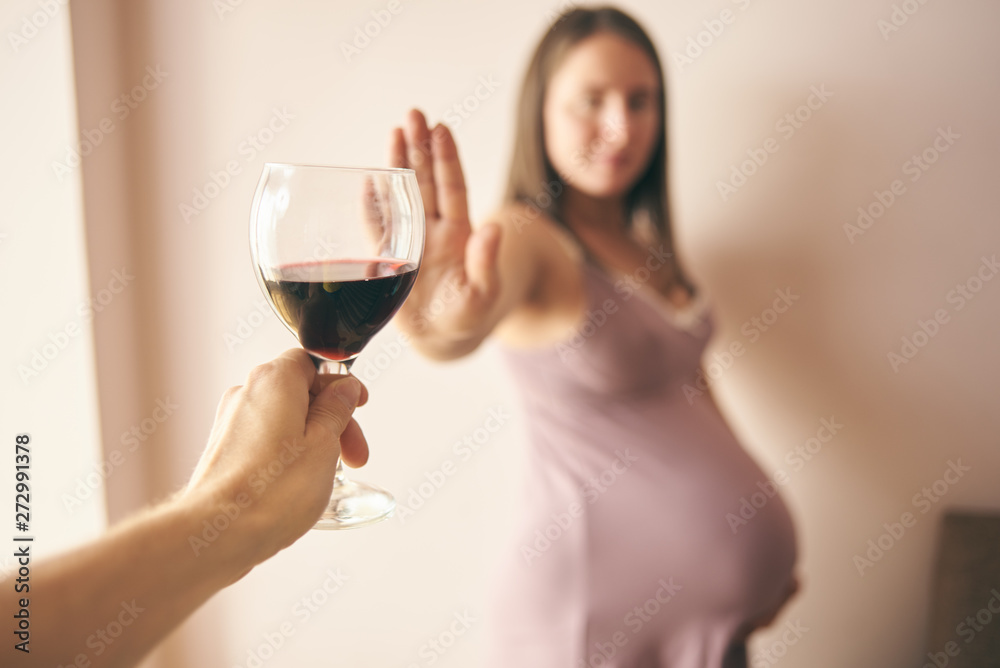 Selective focus of glass of red wine in hand of male and pregnant woman refusing it at background. Future mother leading healthy way of life and caring about health of baby. Concept of pregnancy. Selective focus of glass of red wine in hand of male and pregnant woman refusing it at background. Future mother leading healthy way of life and caring about health of baby. Concept of pregnancy.
