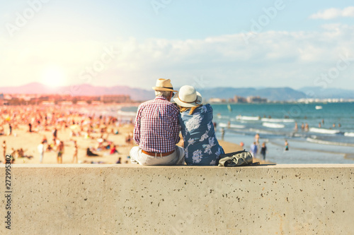 Fototapeta Naklejka Na Ścianę i Meble -  Mature couple of retired lovers enjoying retirement on the beach facing the sea with mobile cell phone taking pictures at sunset. Couple happy true love in the nature