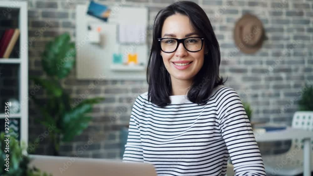Zoom-in portrait of pretty girl wearing glasses and casual clothing using laptop in office then looking at camera smiling. Youth and workplace concept. Zoom-in portrait of pretty girl wearing glasses and casual clothing using laptop in office then looking at camera smiling. Youth and workplace concept.