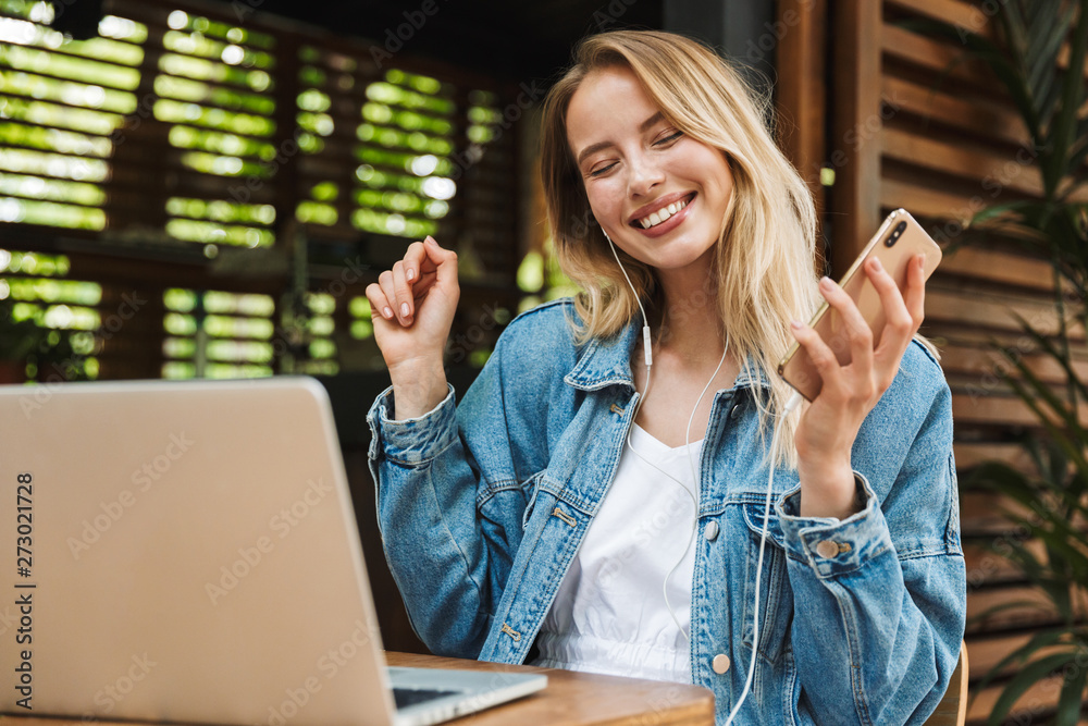 Φωτογραφία Happy young pretty blonde woman in cafe using laptop computer and mobile phone listening music with earphones