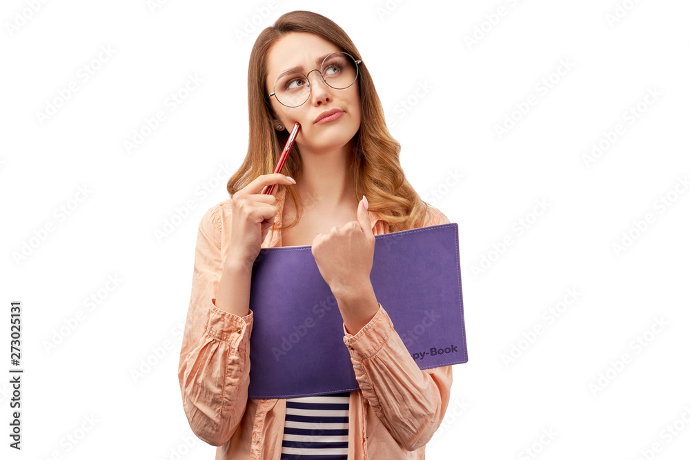 Beautiful young girl has thoughtful expression, holds notepad with pencil, thinks about writing essay, wears optical glasses, isolated over white wall in studio with empty place