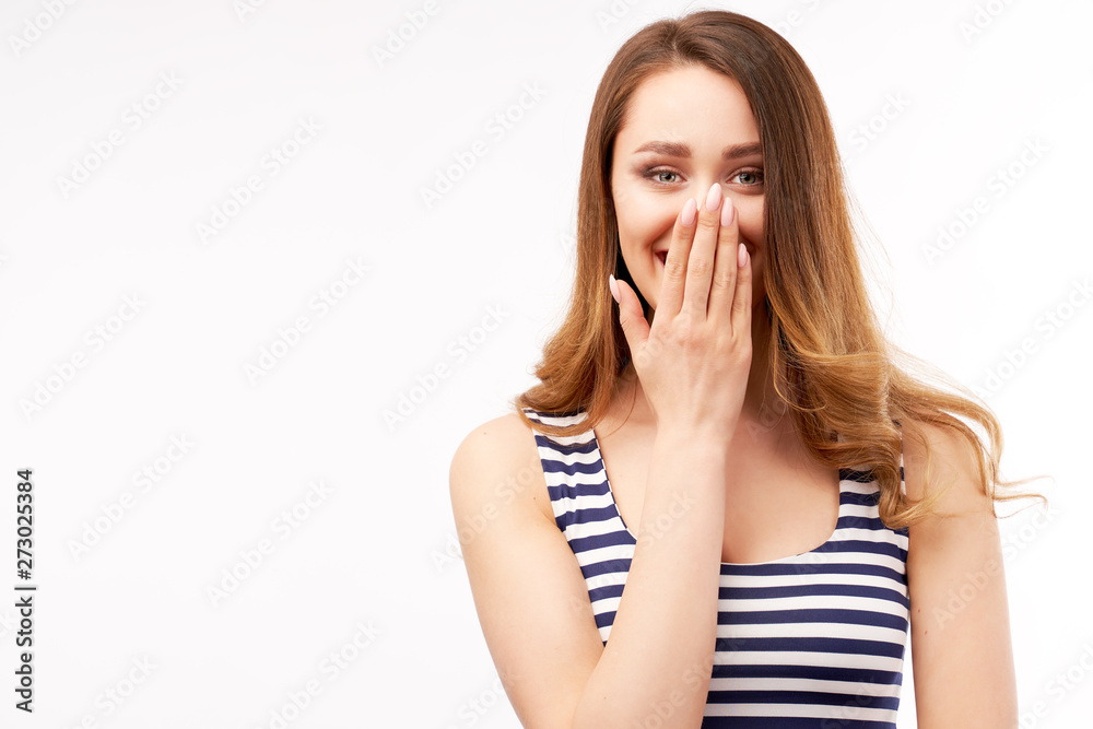 Cheerful glad lovely female with happy expression, covers mouth with hand, smiles positively, dressed in casual striped clothes, isolated on white wall in studio Cheerful glad lovely female with happy expression, covers mouth with hand, smiles positively, dressed in casual striped clothes, isolated on white wall in studio