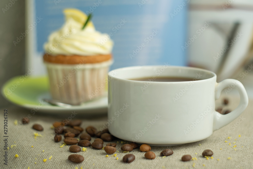 Coffee cup and roasted beans with pastry lemon cupcakes at background with cheese cream, slice of lemon, branch of rosemary at blue background at linen tablecloth, zest, selective focus, close up