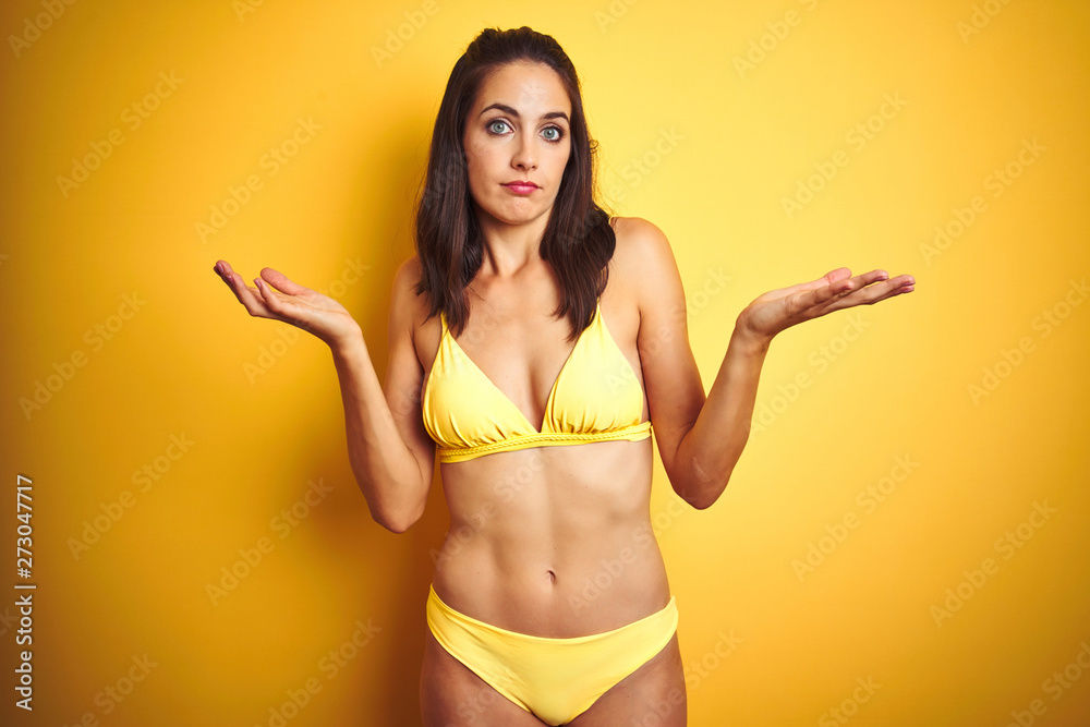 Beautiful woman wearing yellow bikini on summer over isolated yellow  background clueless and confused expression with arms and hands raised.  Doubt concept. Photos | Adobe Stock