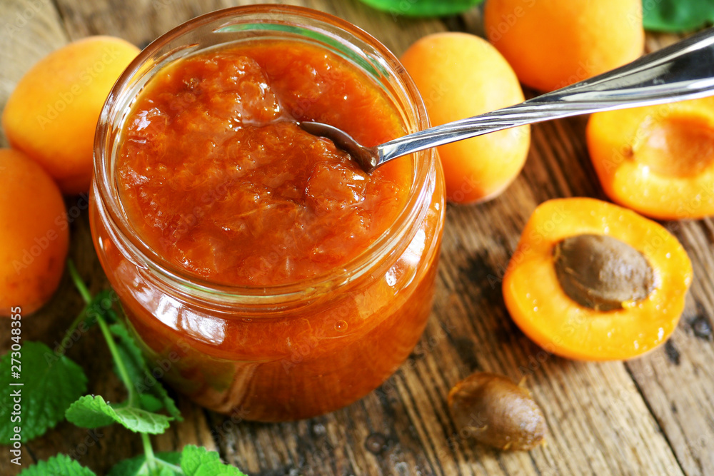 Glass jar of apricot jam on wooden table with ripe apricots at background Glass jar of apricot jam on wooden table with ripe apricots at background