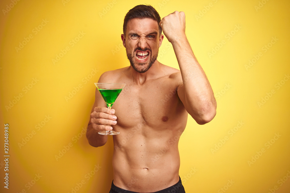 Young handsome shirtless man drinking a summer cocktail over isolated yellow background annoyed and frustrated shouting with anger, crazy and yelling with raised hand, anger concept