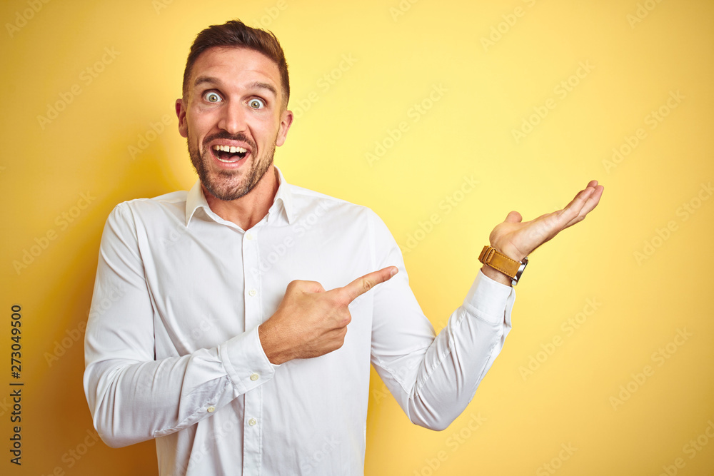 Young handsome man wearing elegant white shirt over yellow isolated background amazed and smiling to the camera while presenting with hand and pointing with finger. Young handsome man wearing elegant white shirt over yellow isolated background amazed and smiling to the camera while presenting with hand and pointing with finger.